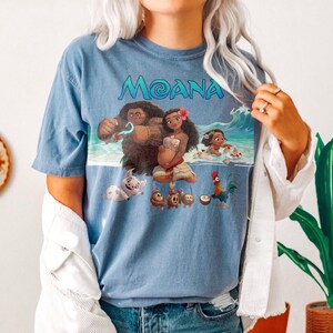 Puede incluir: Camiseta azul con la palabra "MOANA" en escritura turquesa. El gr&aacute;fico representa personajes de la pel&iacute;cula animada, incluyendo a Maui y Moana, en una escena de playa. La camiseta es de color azul liso.