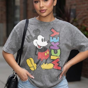 May include: Grey t-shirt with a Mickey Mouse graphic and the word "MICKEY" in rainbow colours. The cartoon character is wearing red shorts and yellow shoes. The shirt has a vintage, washed appearance.