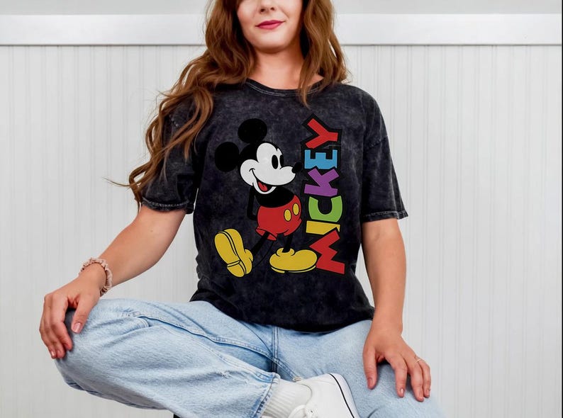 May include: Black t-shirt with a Mickey Mouse graphic. The design shows Mickey in red shorts and yellow shoes, with the word "MICKEY" in rainbow colours. The tee has a vintage, washed appearance.