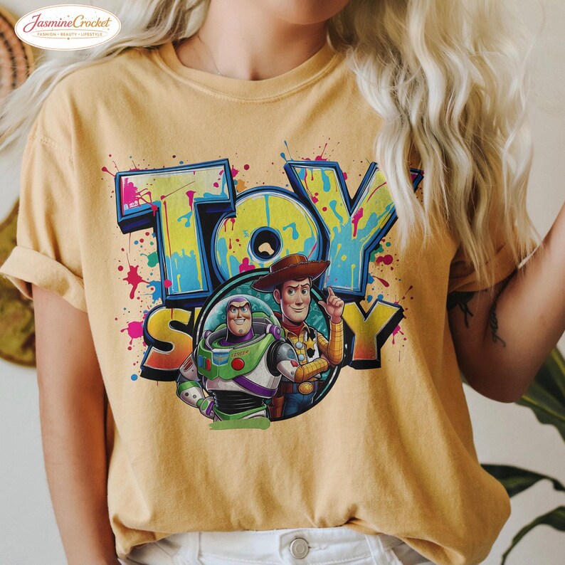 Puede incluir: Camiseta de color amarillo mostaza con el texto "TOY STORY" en un dise&ntilde;o de estilo graffiti colorido. El gr&aacute;fico incluye ilustraciones de Buzz Lightyear y Woody. La camiseta tiene mangas cortas y cuello redondo.