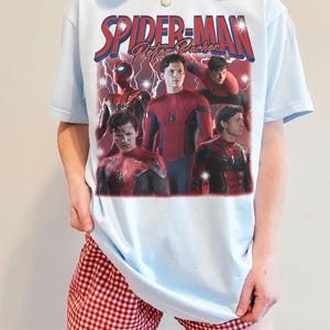 以下が含まれることがあります： 水色のTシャツ。赤と青で「Spider-Man Peter Parker」の文字とスパイダーマンのグラフィックがプリントされています。スパイダーマンの複数の画像が含まれています。モデルは赤と白のチェック柄のショートパンツを着用しています。