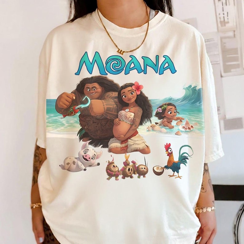 Puede incluir: Camiseta color crema con la palabra "MOANA" en azul, con un gr&aacute;fico de personajes de la pel&iacute;cula animada. El dise&ntilde;o incluye a Maui, Moana y otros personajes en una escena de playa.