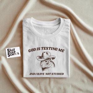 May include: White t-shirt with a humorous graphic featuring a rat wearing a cowboy hat. The text reads "GOD IS TESTING ME AND I HAVE NOT STUDIED". A small "Silly Old School" logo is visible on a card.