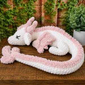 Könnte beinhalten: Ein handgefertigter Plüschdrache in Weiß und Rosa. Der Drache liegt auf einer Holzoberfläche, Kopf und Schwanz ausgestreckt. Der Drache hat rosa Flügel und Akzente sowie schwarz gestickte Augen. Das Gesamtthema ist Fantasy und Kinderspielzeug.