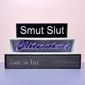 May include: Three-tiered decorative display with the words "Smut Slut", "Cliterature", and "That Sik Luv" in white and purple lettering. The bottom tier also includes the name "JESSIE HALL". The display is set against a pink and purple background.