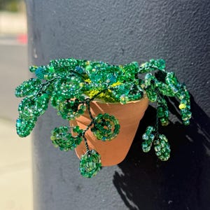 May include: A handcrafted beaded plant in a small terracotta pot. The plant features vibrant green beads, with cascading vines and detailed leaf structures. The pot is attached to a dark gray surface, creating a striking contrast.