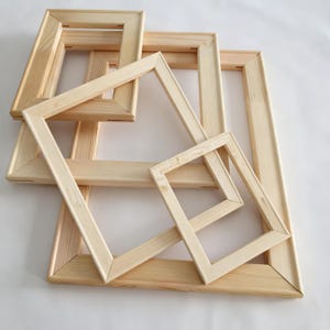 May include: A collection of various sized, unfinished wooden canvas frames. The frames are rectangular and square, with beveled edges and corner joints. The natural wood color is visible, and the frames are arranged on a white surface.