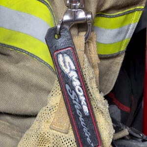 May include: A rectangular black leather tag with a red border and the white text Smoke Showin attached to a silver metal swivel clip. The tag hangs against a tan fabric background featuring horizontal yellow reflective safety strips.