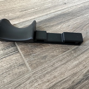 Puede incluir: Una pieza de autom&oacute;vil de pl&aacute;stico negro, posiblemente una moldura o soporte, se muestra sobre una superficie con vetas de madera. La pieza tiene una secci&oacute;n curva y acampanada y una base rectangular con ranuras. El objeto est&aacute; en un entorno neutro.