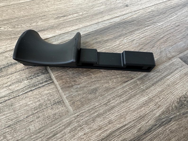 Puede incluir: Una pieza de autom&oacute;vil de pl&aacute;stico negro, posiblemente una moldura o soporte, se muestra sobre una superficie con vetas de madera. La pieza tiene una secci&oacute;n curva y acampanada y una base rectangular con ranuras. El objeto est&aacute; en un entorno neutro.