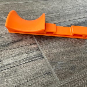 Puede incluir: Herramienta de pl&aacute;stico naranja brillante con una parte superior curva y una base rectangular. La herramienta est&aacute; dise&ntilde;ada con una cuna curva y una base plana y ranurada. El objeto es de pl&aacute;stico naranja opaco.