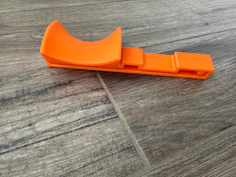 Puede incluir: Herramienta de pl&aacute;stico naranja brillante con una parte superior curva y una base rectangular. La herramienta est&aacute; dise&ntilde;ada con una cuna curva y una base plana y ranurada. El objeto es de pl&aacute;stico naranja opaco.