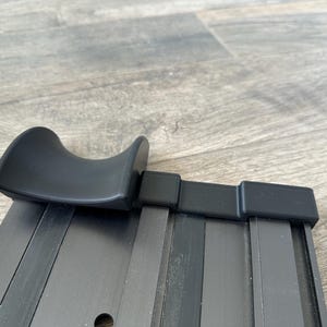 Puede incluir: Una barra de cortina de metal negro con un asa curvada. La barra tiene forma rectangular con ranuras verticales y un acabado liso y mate. El asa tambi&eacute;n es negra y tiene un dise&ntilde;o redondeado y ergon&oacute;mico.