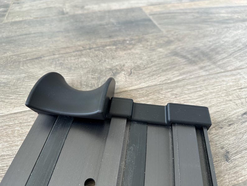 Puede incluir: Una barra de cortina de metal negro con un asa curvada. La barra tiene forma rectangular con ranuras verticales y un acabado liso y mate. El asa tambi&eacute;n es negra y tiene un dise&ntilde;o redondeado y ergon&oacute;mico.