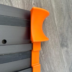 Puede incluir: Primer plano de un componente de pl&aacute;stico naranja fijado a una estructura met&aacute;lica negra. La pieza naranja tiene una parte superior curvada y secciones rectangulares. La estructura met&aacute;lica tiene varias barras negras paralelas.