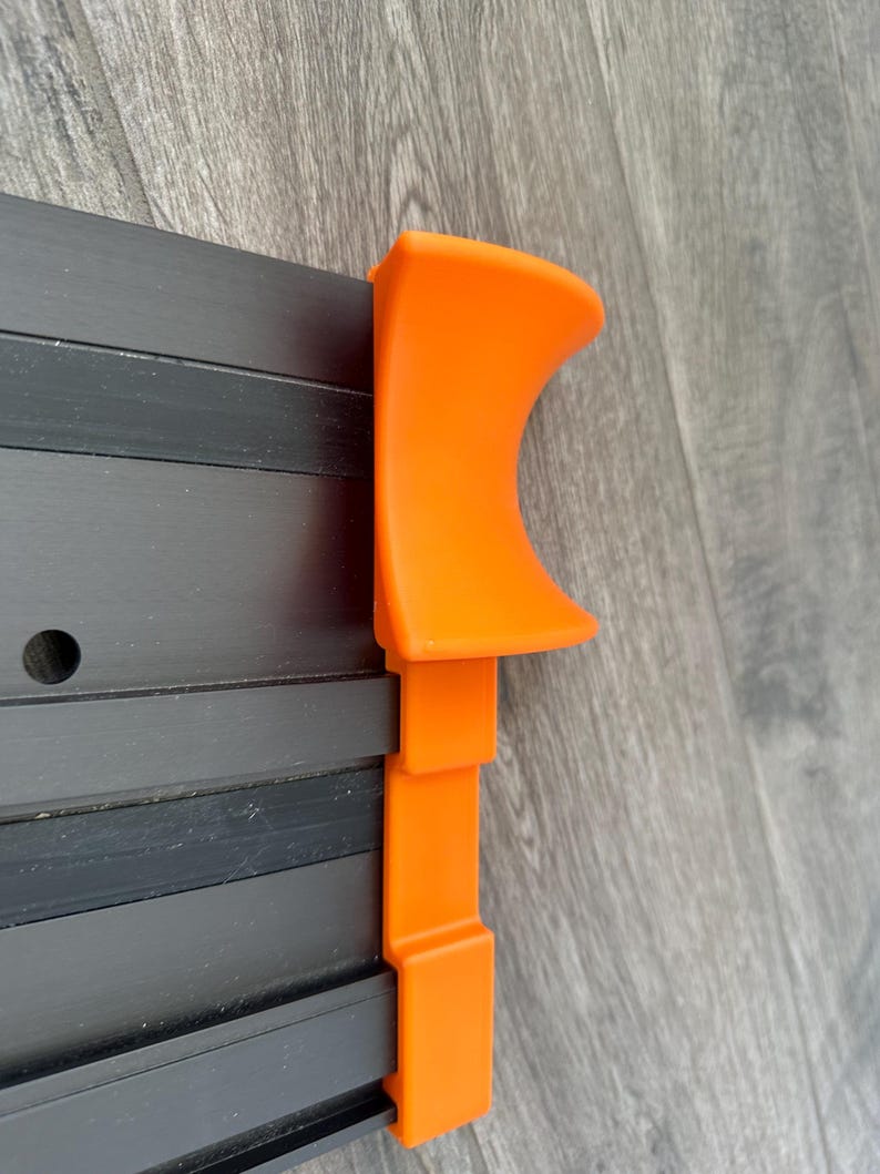 Puede incluir: Primer plano de un componente de pl&aacute;stico naranja fijado a una estructura met&aacute;lica negra. La pieza naranja tiene una parte superior curvada y secciones rectangulares. La estructura met&aacute;lica tiene varias barras negras paralelas.