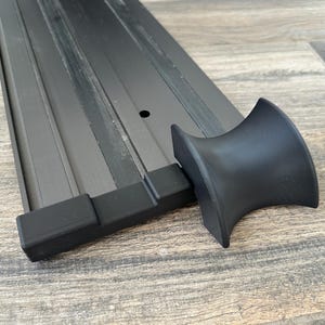 Puede incluir: Un objeto met&aacute;lico negro con un dise&ntilde;o curvo, posiblemente un componente de mueble o una pieza decorativa. El objeto se encuentra sobre una superficie de madera, resaltando su forma y textura. El dise&ntilde;o presenta una serie de l&iacute;neas paralelas.