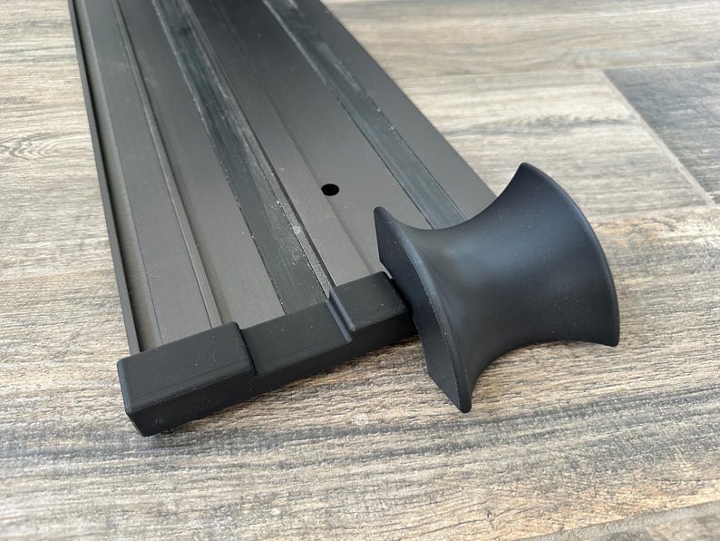 Puede incluir: Un objeto met&aacute;lico negro con un dise&ntilde;o curvo, posiblemente un componente de mueble o una pieza decorativa. El objeto se encuentra sobre una superficie de madera, resaltando su forma y textura. El dise&ntilde;o presenta una serie de l&iacute;neas paralelas.
