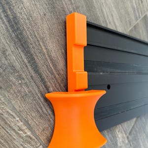 Puede incluir: Una herramienta de pl&aacute;stico naranja con una base acampanada y un poste vertical con una parte superior rectangular. La herramienta est&aacute; montada en un riel de metal negro con ranuras horizontales. El fondo es una superficie de vetas de madera.