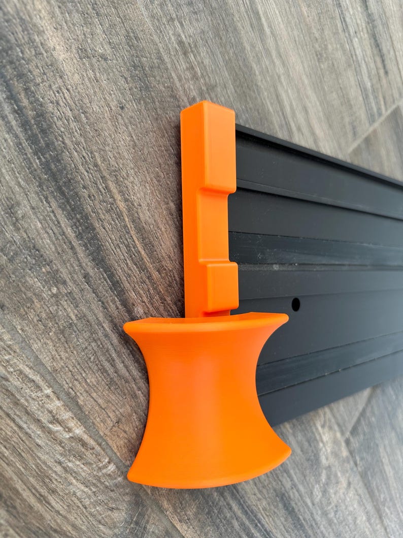 Puede incluir: Una herramienta de pl&aacute;stico naranja con una base acampanada y un poste vertical con una parte superior rectangular. La herramienta est&aacute; montada en un riel de metal negro con ranuras horizontales. El fondo es una superficie de vetas de madera.