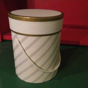 May include: A white cylindrical gift box with a gold trim and a diagonal gray and white striped pattern. The box has a white rope handle.