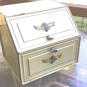 May include: A vintage cream-colored metal bread box with a slanted hinged lid and a lower drop-down door. Both the lid and the door feature a colorful geometric decal with red, blue, yellow, and green shapes. A small blue knob is on the lower door.