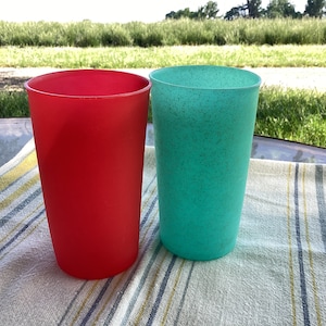 May include: Two tall plastic tumblers, one red and one teal, sit on a striped cloth. The red tumbler has a solid color, while the teal one has a speckled texture. The background shows a grassy area.