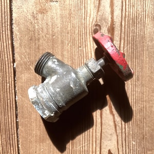 May include: A vintage, weathered metal valve with a red handle. The valve is a dull silver color with signs of age and use. The red handle shows wear, with paint chipping off. The valve is set against a wooden background.