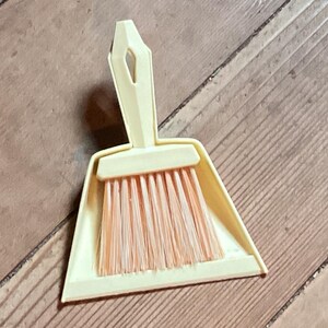 May include: A small, cream-colored dustpan and brush set. The dustpan is triangular with a handle, and the brush has peach-colored bristles. The set is made of plastic and is designed for small cleaning tasks.
