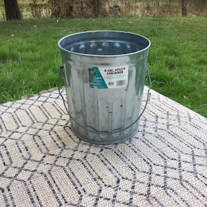 May include: A silver 6-gallon utility container with a black handle. The container is sitting on a white and black patterned rug.