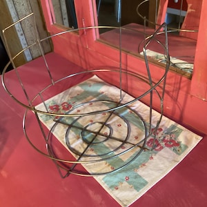 May include: A vintage metal wire basket with a circular design, resting on a floral patterned cloth. The basket has a silver finish and is set against a red surface, with a red-painted wooden frame in the background.