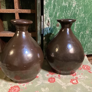 May include: Two dark brown ceramic vases with a rounded body and a narrow neck. The vases are sitting on a floral patterned tablecloth.