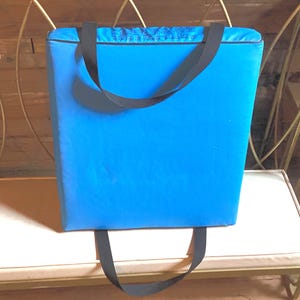 May include: A bright blue tote bag with black handles. The bag is rectangular and appears to be made of a sturdy material. The handles are long enough to be carried over the shoulder. The bag is sitting on a white bench.