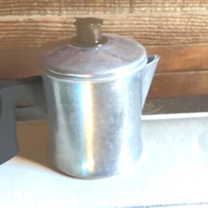 May include: A silver-colored metal percolator coffee pot with a cylindrical body, a short spout, and a dark brown handle. The pot features a matching lid with a dark brown knob on top. The surface of the metal has a slightly textured, matte finish.