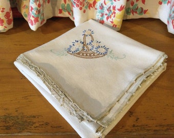 May include: A white linen tablecloth with a delicate embroidered design of a basket of flowers. The tablecloth has a scalloped edge and is folded in half.