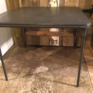 May include: A dark gray folding table with a rectangular top and metal legs. The table is supported by a wooden brace and is set against a rustic wooden wall. The table is approximately 30 inches long.