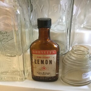 May include: A brown glass bottle with a black cap, labeled "Burnett's Pure Extract Lemon". The label also includes the text "Alcohol 85 Percent, Two Ounces Net" and "Mfd by Joseph Burnett Co., Boston Mass."