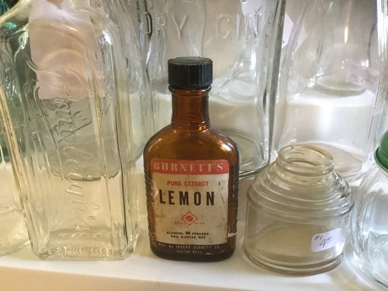 May include: A brown glass bottle with a black cap, labeled "Burnett's Pure Extract Lemon". The label also includes the text "Alcohol 85 Percent, Two Ounces Net" and "Mfd by Joseph Burnett Co., Boston Mass."
