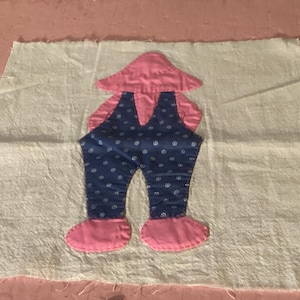 May include: A handmade quilt block featuring a whimsical figure wearing blue overalls with white polka dots and pink shoes. The block is made of cotton fabric and is set against a white background.
