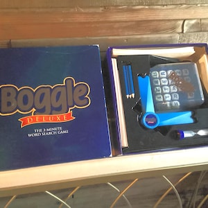 May include: A Boggle Deluxe word search game box, open to reveal the game components. The box is blue with the game title in gold lettering. Inside are pencils, a timer, and a letter grid.