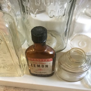 May include: A collection of vintage glass bottles, including a clear glass bottle with the text "LONDON DRY GIN", a clear glass bottle with the text "Distilled in the USA", a clear glass bottle with a wide mouth, and a brown glass bottle with a black cap and the text "BURNETT'S PURE EXTRACT LEMON".