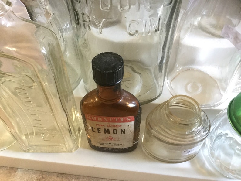 May include: A collection of vintage glass bottles, including a clear glass bottle with the text "LONDON DRY GIN", a clear glass bottle with the text "Distilled in the USA", a clear glass bottle with a wide mouth, and a brown glass bottle with a black cap and the text "BURNETT'S PURE EXTRACT LEMON".