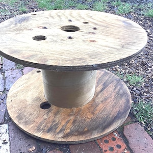 May include: A rustic, round wooden table made from a repurposed cable spool. The table has a light brown top with several holes and a darker brown base. The central support is a light brown cylinder. The table is outdoors.