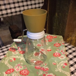 May include: A vintage hand-crank food grinder with a clear glass jar and a yellow plastic funnel. The grinder has a white plastic handle and a metal crank.