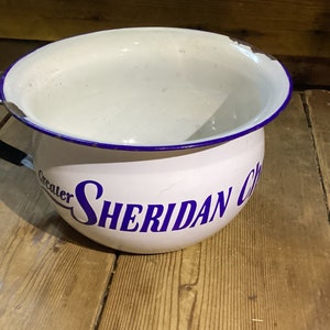 May include: White enamel chamber pot with a blue rim and a black handle. The words "Greater Sheridan Club" are printed in blue on the side. The pot sits on a wooden surface.