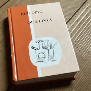 May include: A hardcover book titled "BUILDING OUR LIVES" with an orange spine and a light pink cover. The cover features a line drawing of tools and a box. The book is resting on a wooden surface.