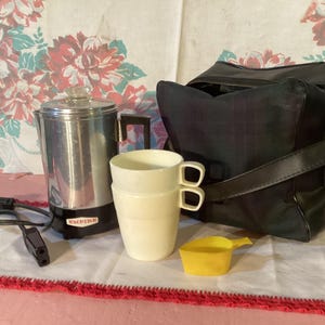 May include: A silver electric coffee percolator with a black base and the word "Empire" printed on it. The percolator is on a white tablecloth with a red trim. There are two white ceramic mugs and a yellow measuring spoon on the table. A black leather bag is in the background.