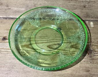 May include: A green glass saucer with a decorative pattern. The saucer has a scalloped edge and a raised center.