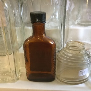 May include: A collection of five vintage glass bottles, including a brown glass bottle with a black lid, a clear glass bottle with a textured surface, and a clear glass bottle with a ribbed surface.
