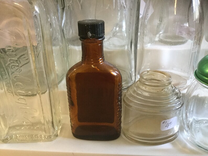 May include: A collection of five vintage glass bottles, including a brown glass bottle with a black lid, a clear glass bottle with a textured surface, and a clear glass bottle with a ribbed surface.